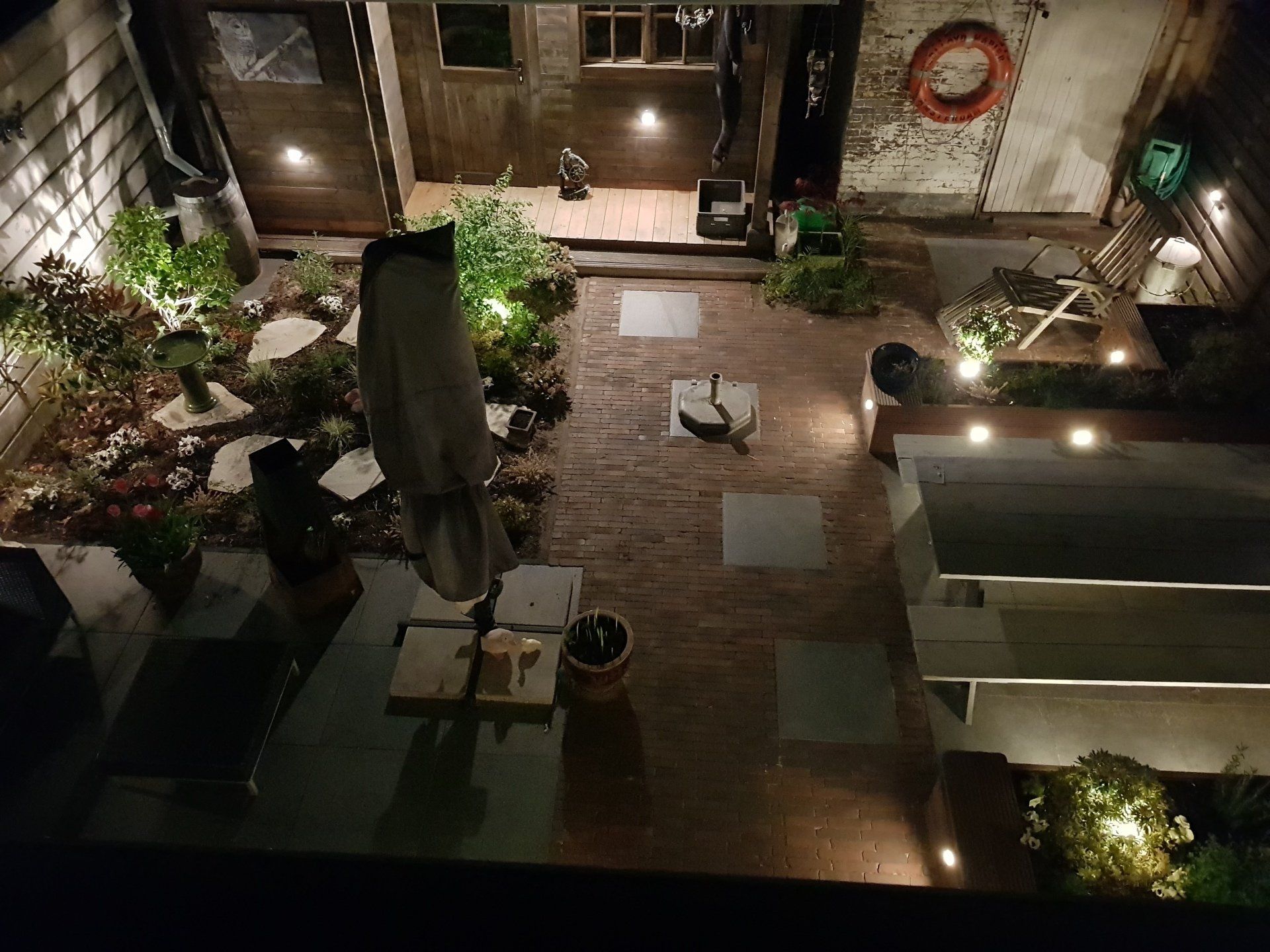 Nachtelijk uitzicht op een bakstenen binnenplaats met stenen bestrating, verlicht door tuinverlichting. Planten en meubilair zijn zichtbaar.