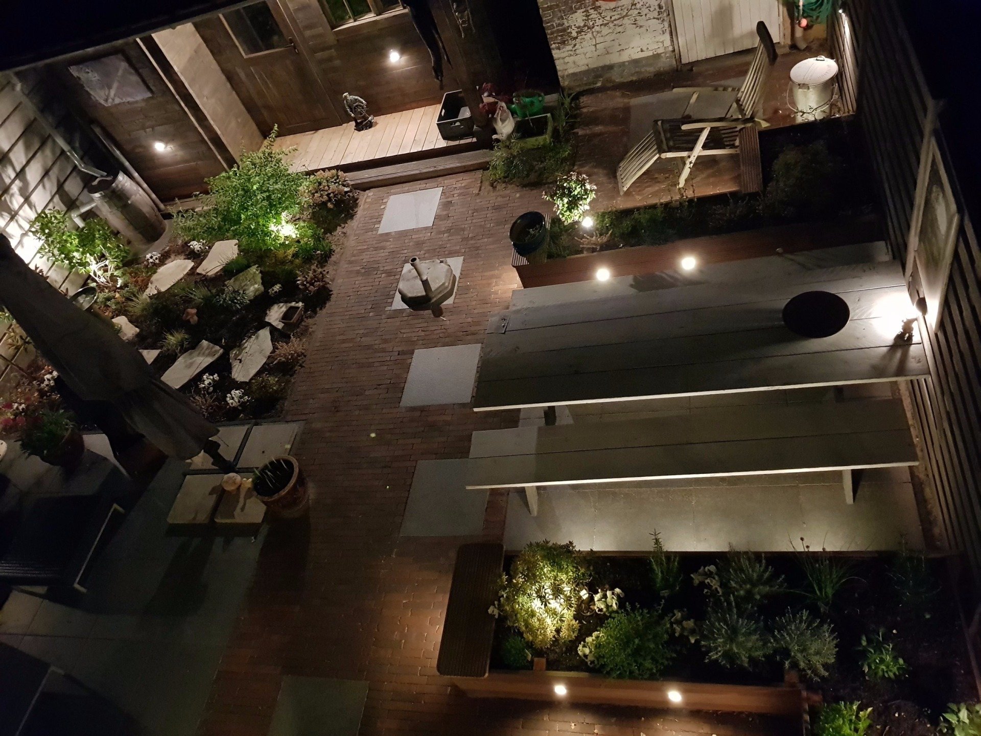Bovenaanzicht van een verlicht terras 's nachts. Bakstenen en stenen vloer, groen en ingebouwde verlichting zijn te zien.