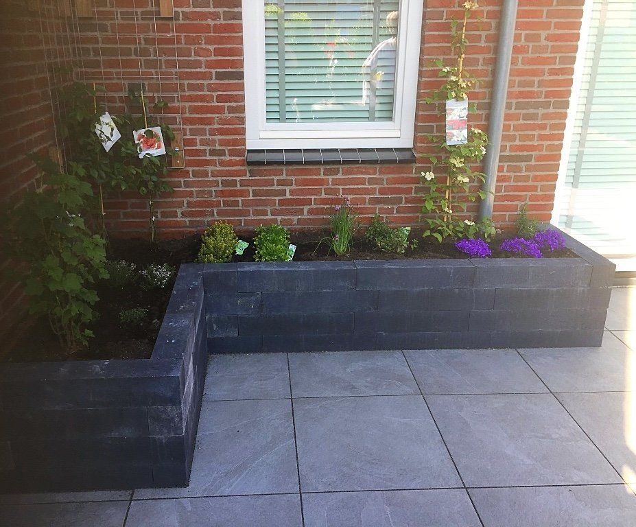 Rechthoekig, verhoogd tuinbed met planten tegen een bakstenen muur. Grijs betegeld terras ervoor.