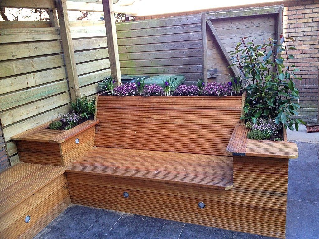 Houten buitenbank met ingebouwde plantenbakken, omgeven door een houten schutting en een bakstenen muur.