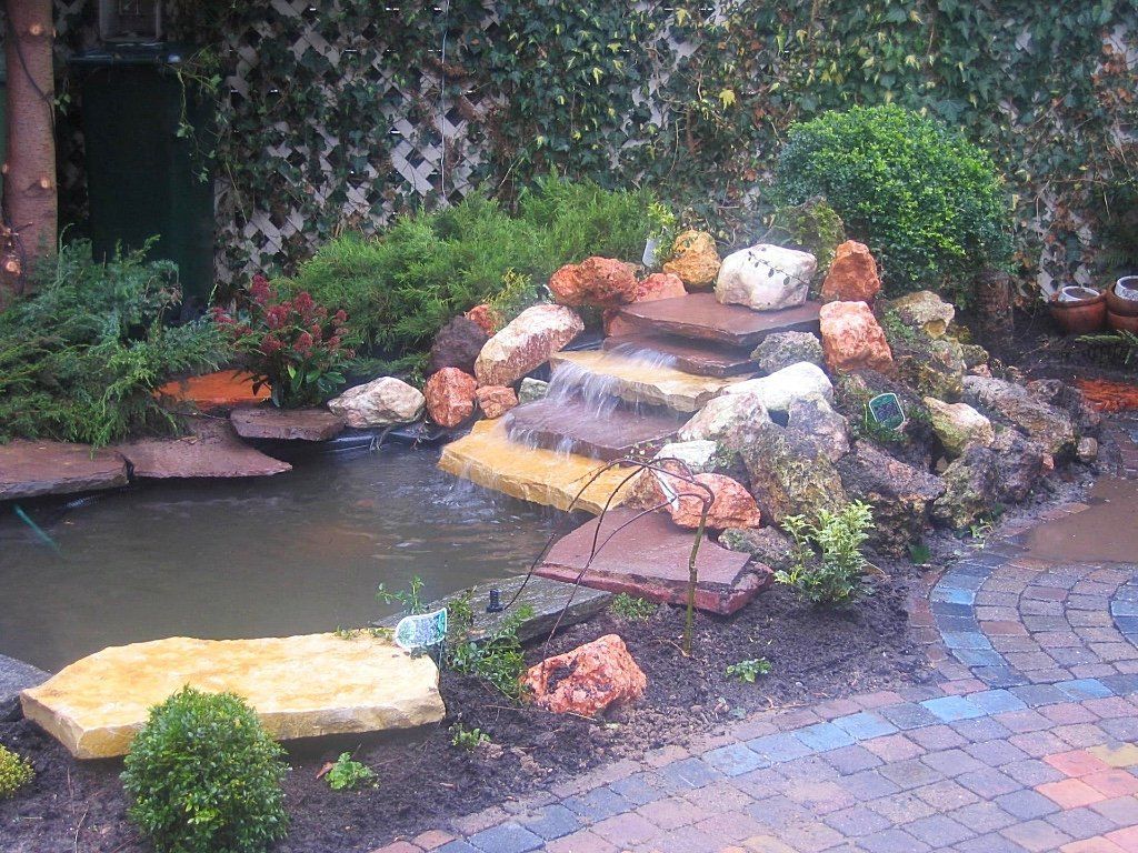 Water stroomt via een rotswaterval naar beneden in een vijver, omgeven door groen en bestrating.