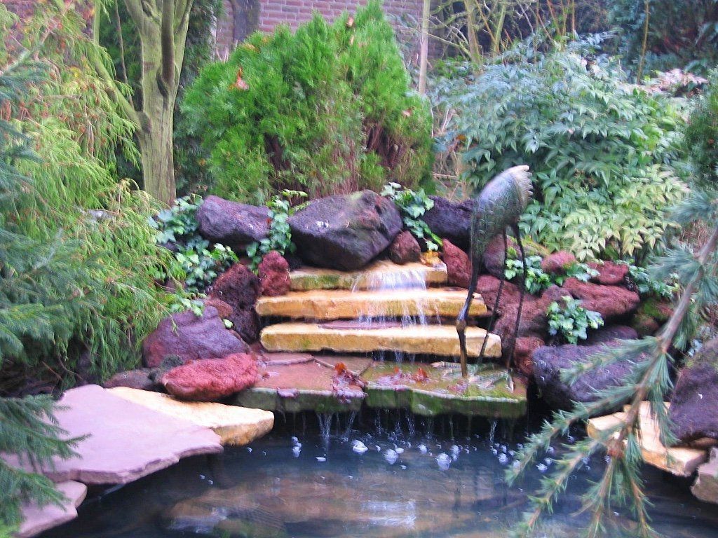 Water stroomt via stenen trappen naar beneden in een kleine vijver, omgeven door weelderig groen en rotsen.