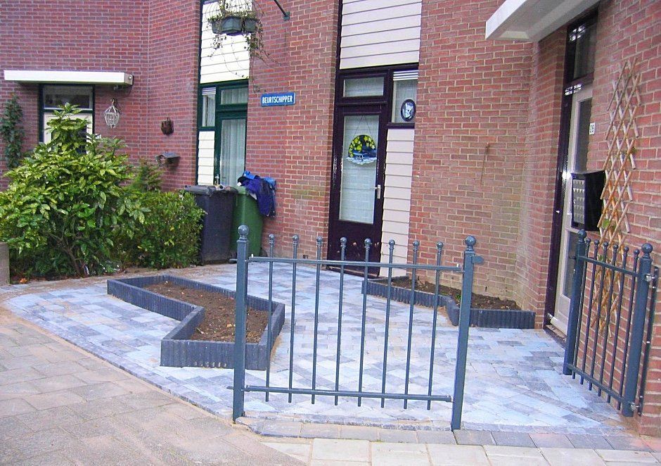 Bakstenen gebouwen, kleine tuin, grijs metalen hek, bestraat pad.