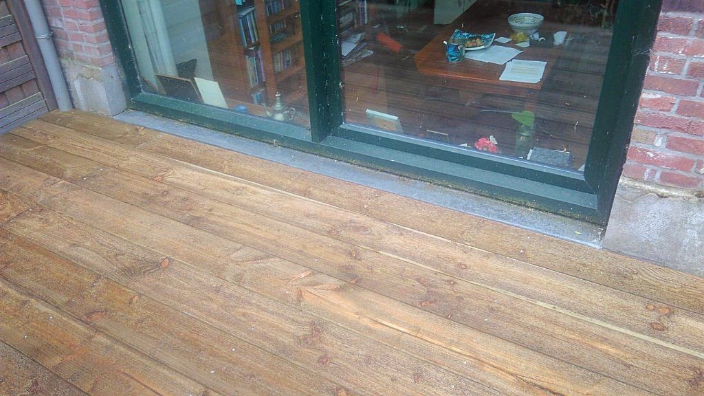 Houten terras voor een raam, grenzend aan een bakstenen muur. In het raam worden een tafel en stoelen weerspiegeld.