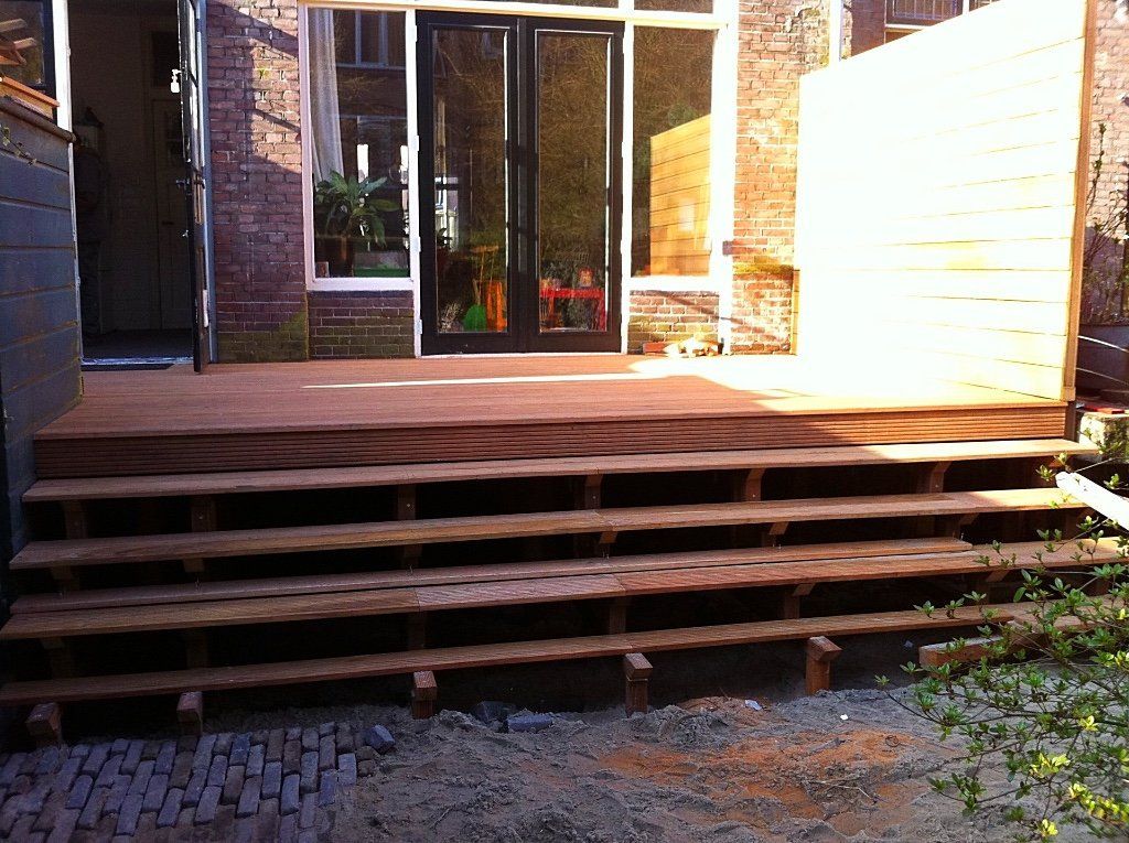 Houten terras met meerdere treden die leiden naar glazen deuren van een bakstenen gebouw.