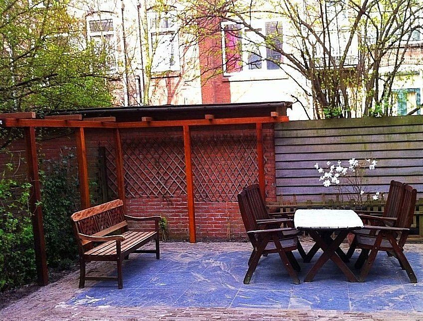 Terras met houten meubilair: bank, tafel, stoelen en een pergola in de achtertuin.