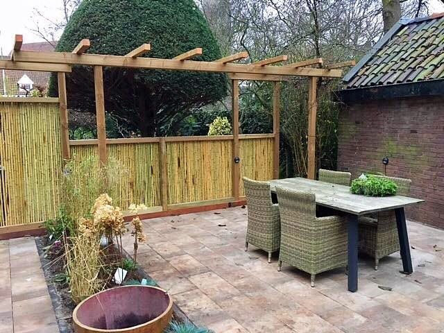 Terras met houten pergola, eettafel, rieten stoelen, bamboeschutting en beplanting.