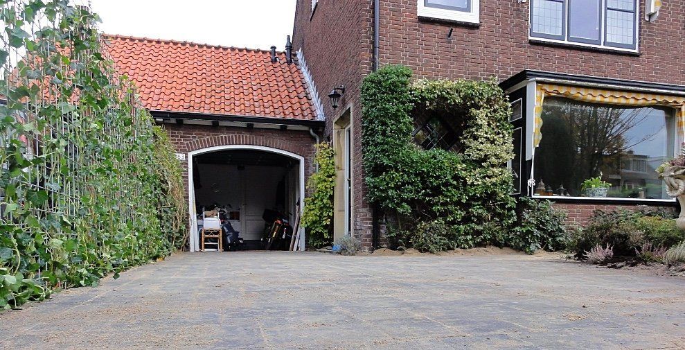 Oprit die leidt naar een bakstenen huis met een garage en een raam omgeven door groen.