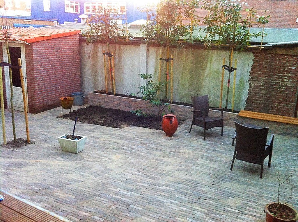 Een klein bakstenen terras met stoelen, bomen en potplanten tegen een betonnen muur.