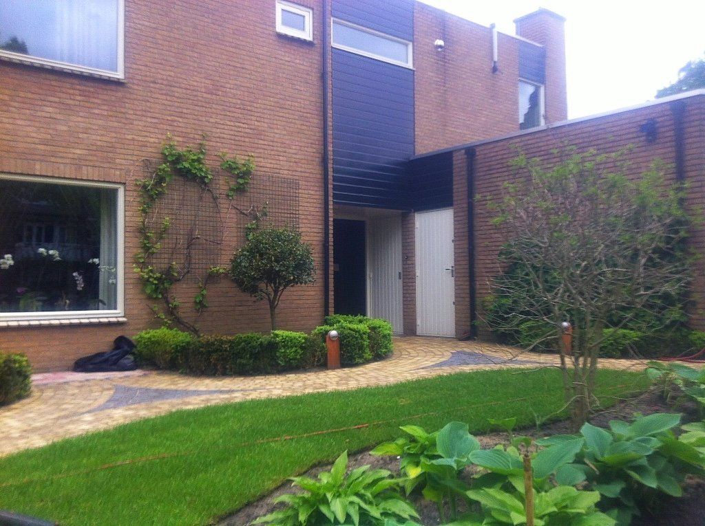 Een bakstenen huis met een aangelegde tuin; een pad leidt naar een terugliggende ingang onder een donkere gevel.