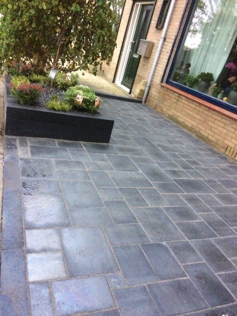 Een donkergrijze, geplaveide stoep met een verhoogde, zwarte plantenbak gevuld met bloemen naast een gebouw.