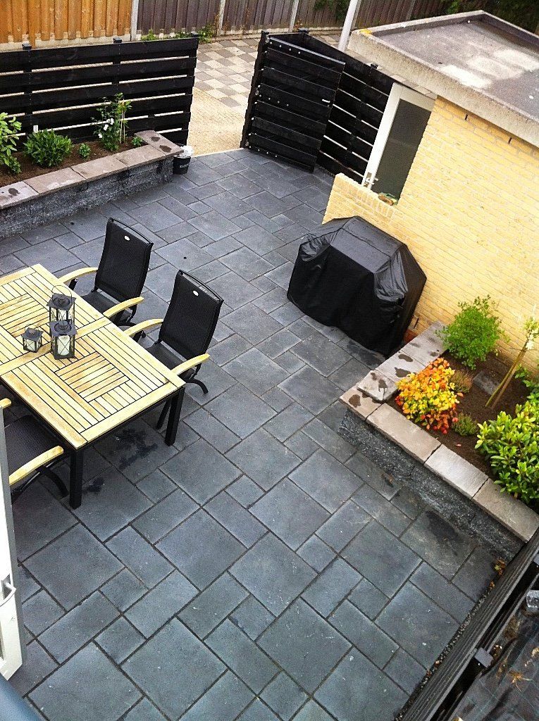 Terras met grijze natuurstenen tegels, houten tafel met stoelen, zwarte barbecue en lage plantenbakken met bloemen.
