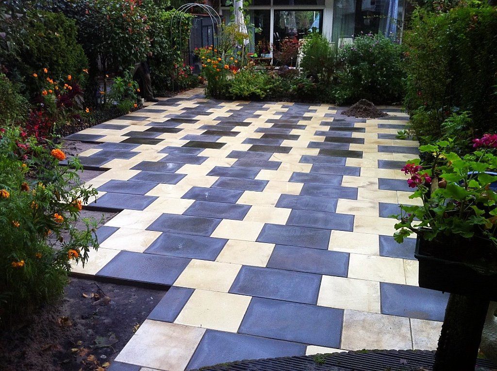 Buitenpatio bestraat met afwisselend blauwe en crèmekleurige tegels, omgeven door tuinbeplanting.