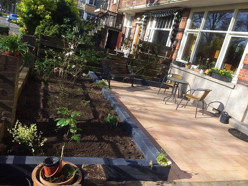 Een bloembed met pas omgewoelde aarde, grenzend aan een terras met zitgelegenheid bij een bakstenen gebouw.