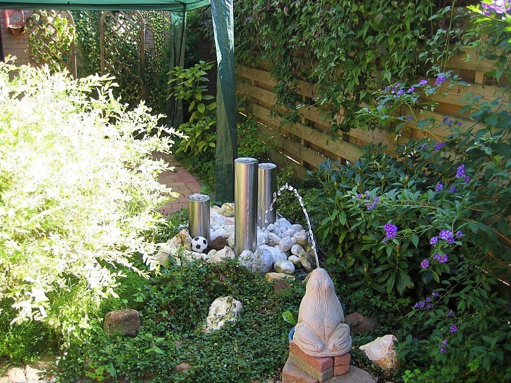 Een kleine tuin met een fontein van zilverkleurige buizen, omgeven door groen en rotsen. Er staat een kikkerbeeld voor.