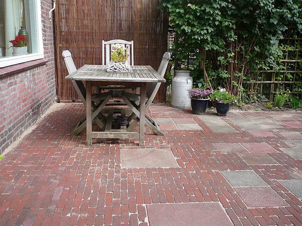 Een stenen terras met een houten tafel en stoelen, vlakbij een bakstenen muur en begroeiing.