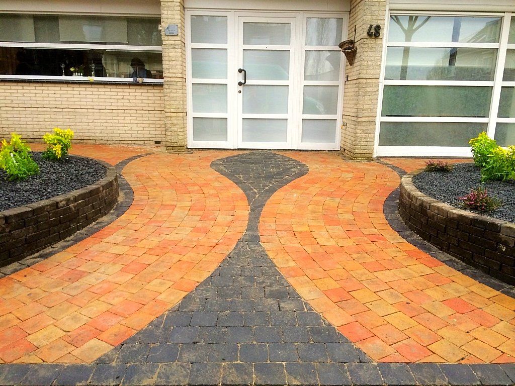 Een bakstenen pad leidt naar een witte deur, omlijst door lage bloembedden en een garagedeur.