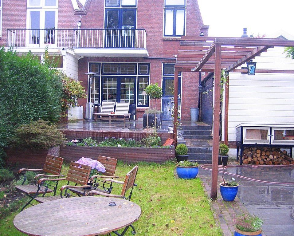 Achtertuin met bakstenen huis, terras, houten pergola, gazon en zitgelegenheid buiten.