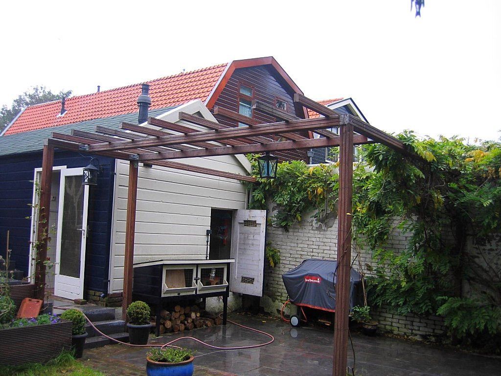 Bruine pergola boven een wit-blauwe schuur met rood dak, in een achtertuin.