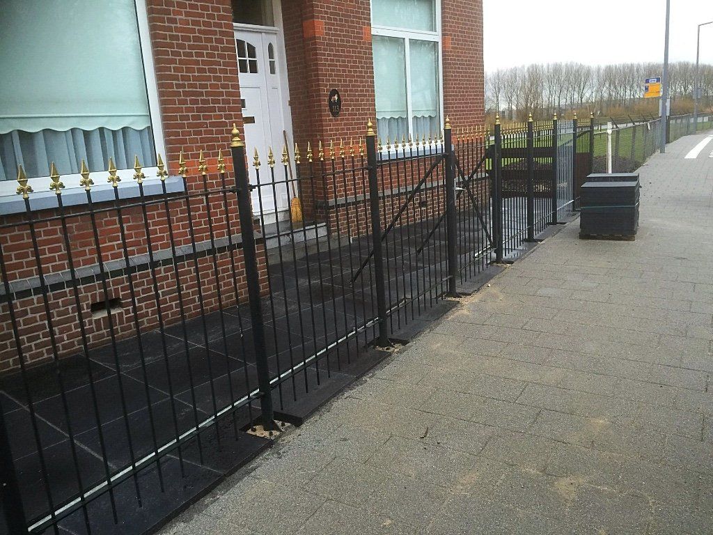 Zwart ijzeren hek met gouden sierelementen, bakstenen gebouw en trottoir.