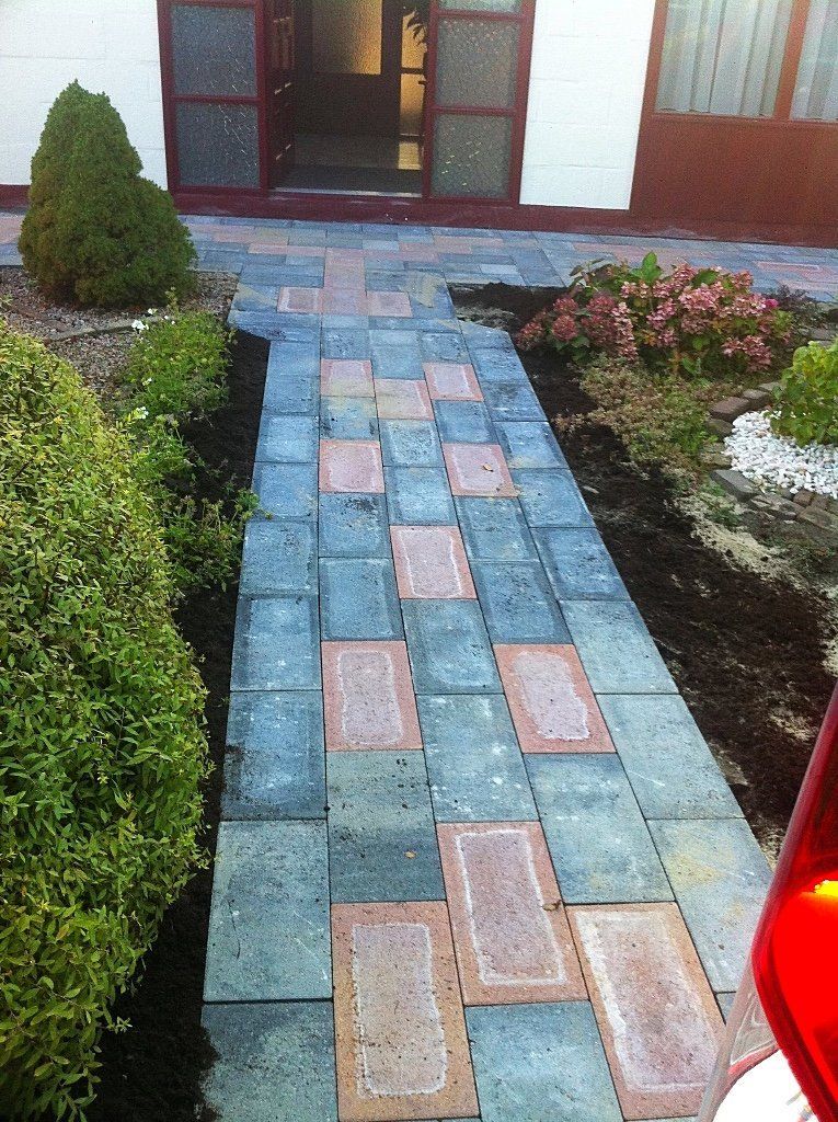 Een bakstenen pad leidt naar een deuropening, geflankeerd door groene struiken en bloemperken, met contrasterende blauwe en rode bakstenen.