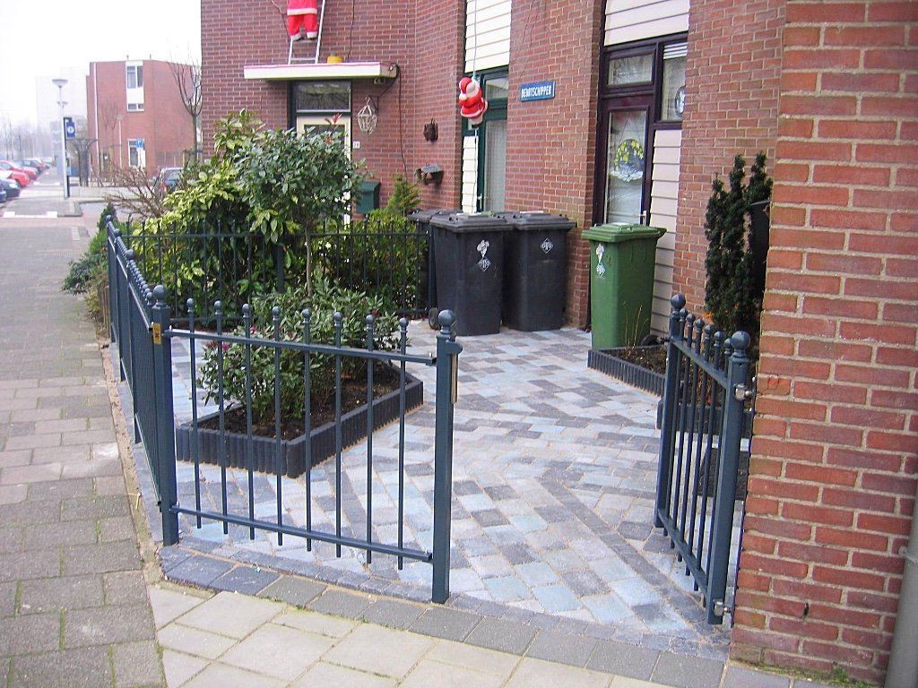 Buitenkant van bakstenen gebouwen met een omheind gedeelte van bestrating met struiken en afvalbakken naast een trottoir.