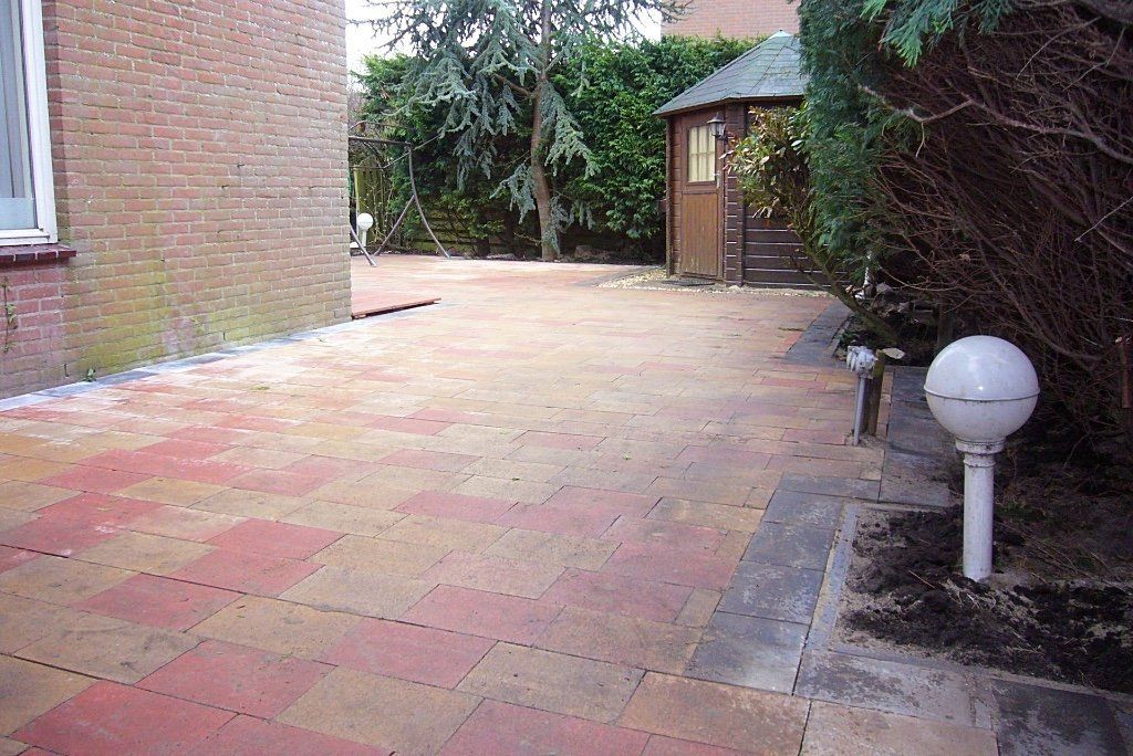Een stenen terras met oranje en gele tegels leidt naar een schuur, omgeven door struiken en een stenen gebouw.