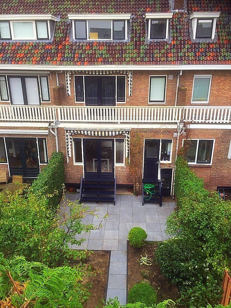 Een bakstenen gebouw met meerdere ramen en een balkon met uitzicht op een tuin met een stenen pad en groen.