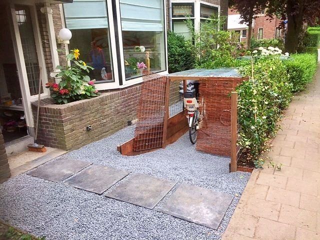De fiets staat beschut onder een constructie van baksteen en hout naast een huis met een ondergrond van grind en betontegels.