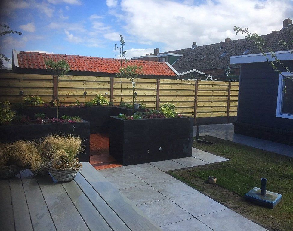 Tuin met zwarte plantenbakken, houten schutting, gras en een terras op een bewolkte dag.