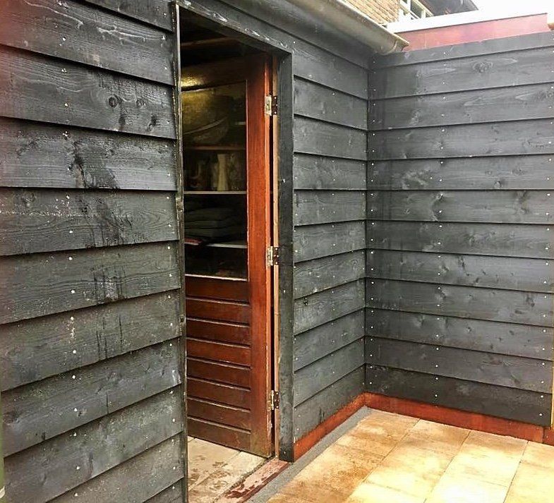 Zwarte houten schuur met open bruine deur, waardoor de schappen aan de binnenkant zichtbaar zijn.