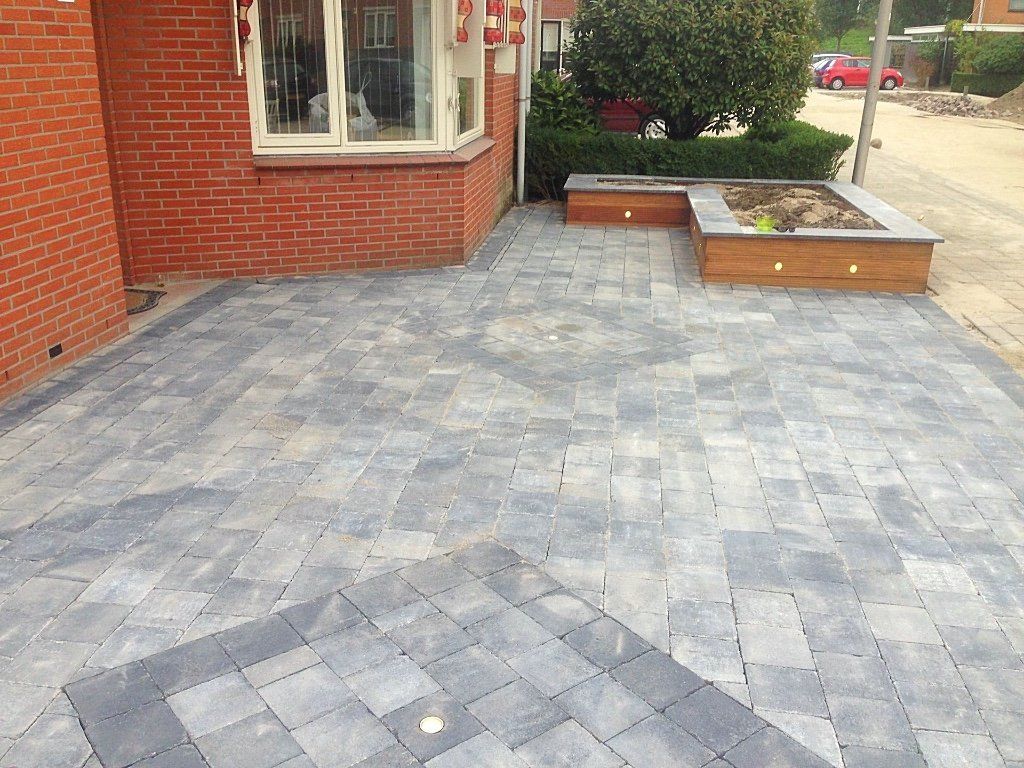 Bakstenen huis met een betegeld terras. Grijze en donkergrijze bestrating. Houten plantenbakken gevuld met groen.
