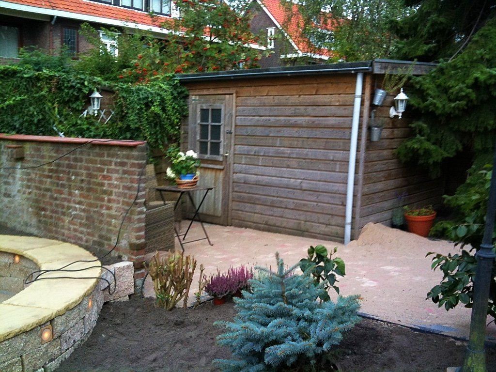 Houten schuur in een tuin met een bakstenen muur, grindondergrond en groen.