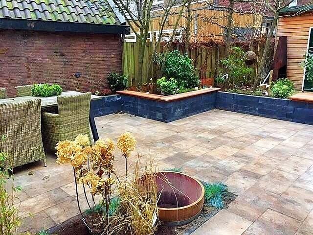 Terras met bakstenen muur, verhoogde bloembedden, rieten tuinmeubelen en bruine bestrating.