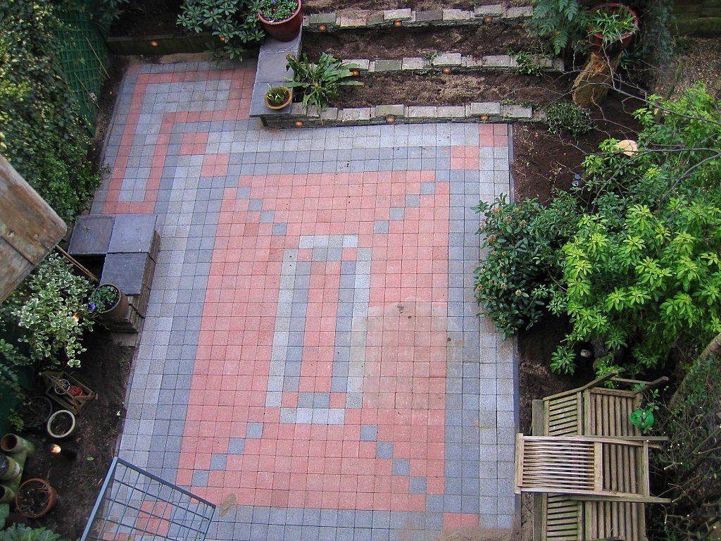 Bovenaanzicht van een bakstenen terras met rode en grijze bestrating, bloembedden en groen.