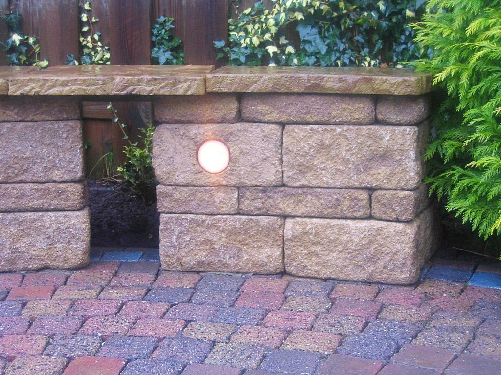 Stenen muur met ingebouwde verlichting, bakstenen terras en groen op de achtergrond.