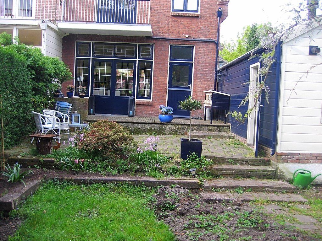 Achtertuin met een bakstenen huis, een trap en een kleine tuin met planten. Blauwe deuren en witte stoelen zijn zichtbaar.