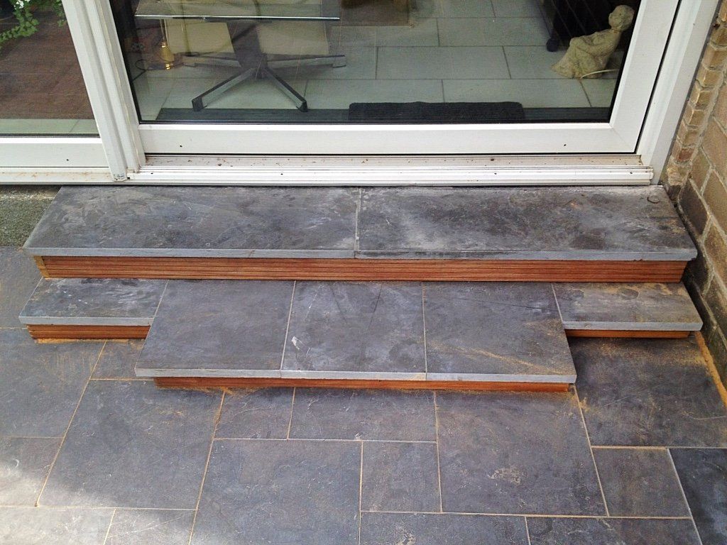 Stenen trappen leiden naar een schuifdeur van glas. Donkergrijze tegels en houten afwerking.