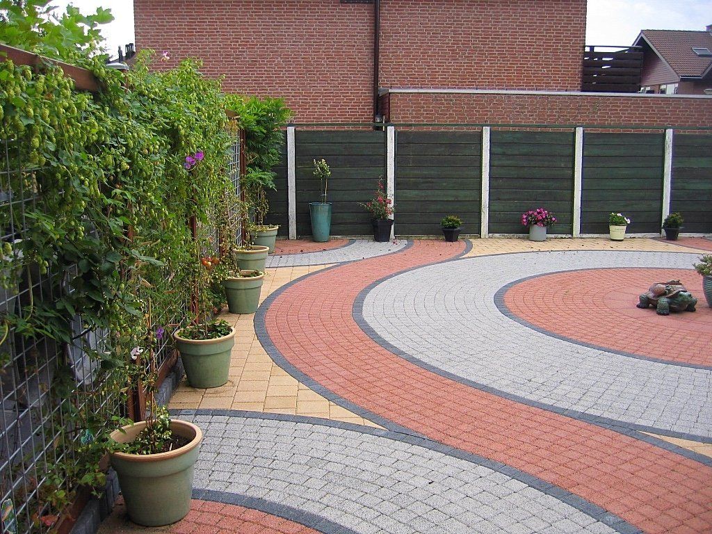 Een betegeld terras met concentrische cirkels in rood, grijs en geel. Groene potplanten staan ​​langs een met klimplanten begroeide schutting.