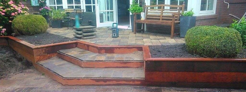 Een trap leidt naar een patio met bloemperken, banken en een gebouw met witte deuren.