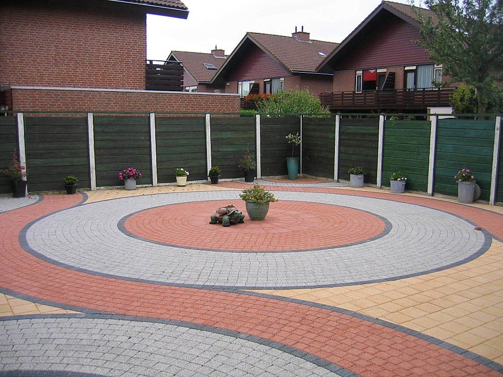 Rond bakstenen terras met bloempotten, omgeven door een groene schutting en huizen op de achtergrond.