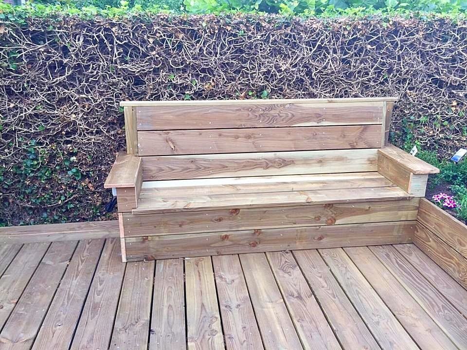 Houten buitenbank op een houten terras, tegen een groene heg aan.