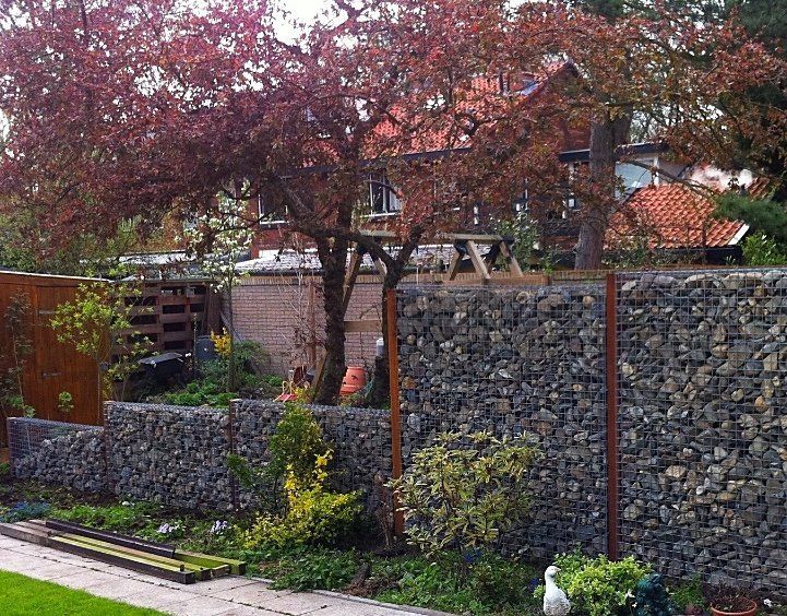 Gabionmuren met een boom met roodachtige bladeren ervoor, naast een roestig metalen hek en een huis met een pannendak.