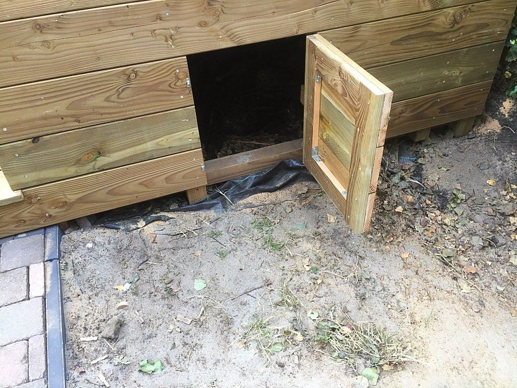Houten hondenhok met open deur, geplaatst op een ondergrond van zand en aarde.