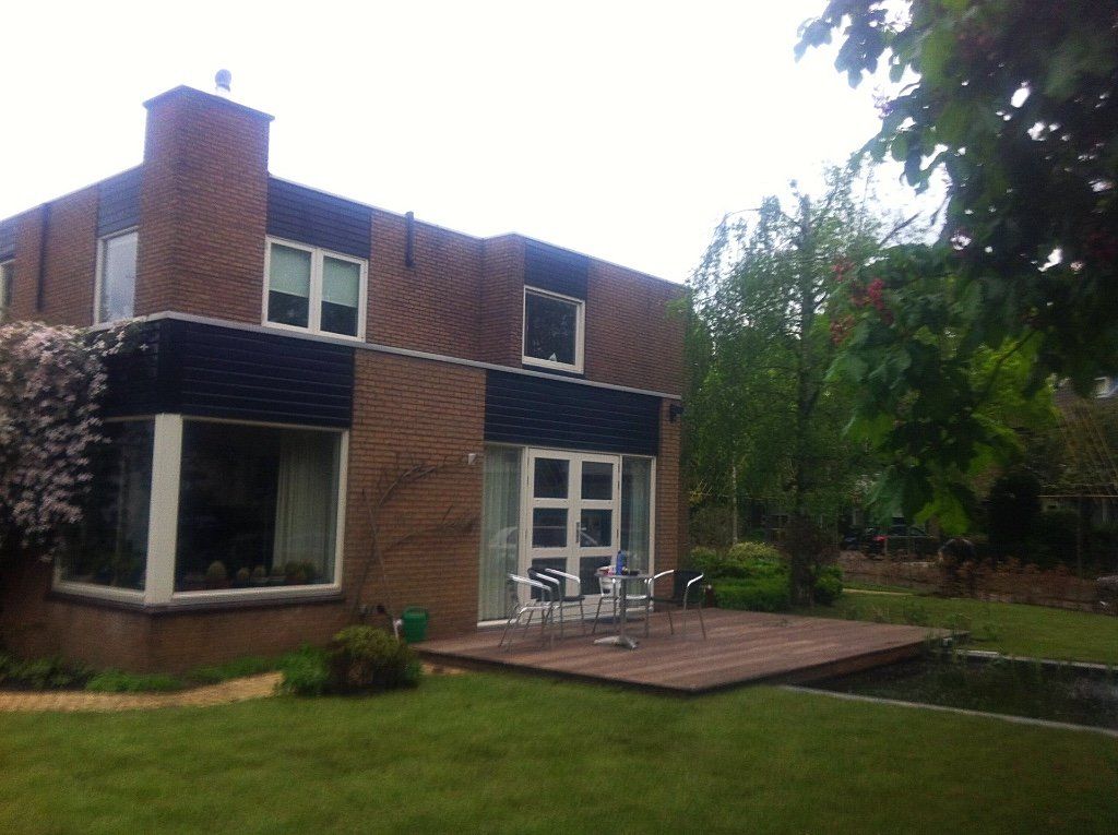 Een bakstenen huis met zwarte kozijnen, een terras met stoelen en een groen gazon.
