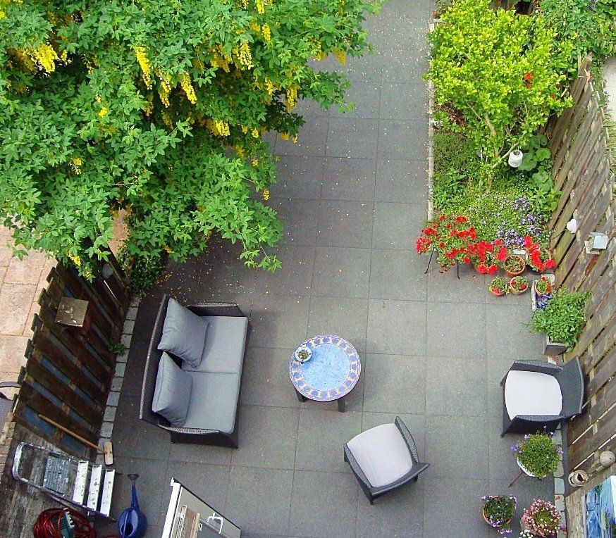 Bovenaanzicht van een terras met grijs meubilair, een kleine ronde tafel en diverse potplanten rondom een ​​betegeld gedeelte.