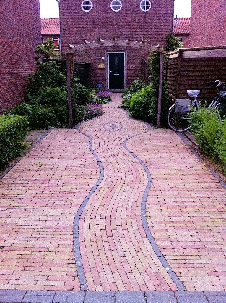 Een bakstenen pad met een gebogen grijze rand leidt naar de ingang van een bakstenen gebouw. ​​Langs het pad staan ​​weelderige groene planten.