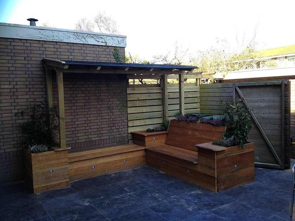 Een zitgedeelte buiten met houten banken, plantenbakken en een kleine overkapping tegen een bakstenen muur en houten schutting.