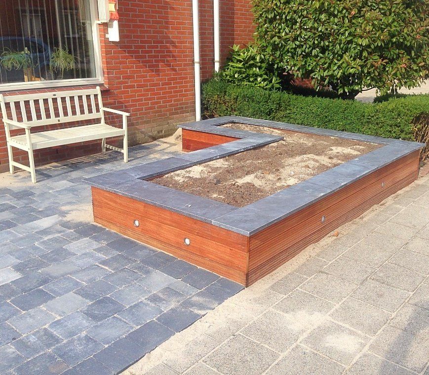 Houten zandbak met een donkere rand, naast een witte bank en een bakstenen gebouw.