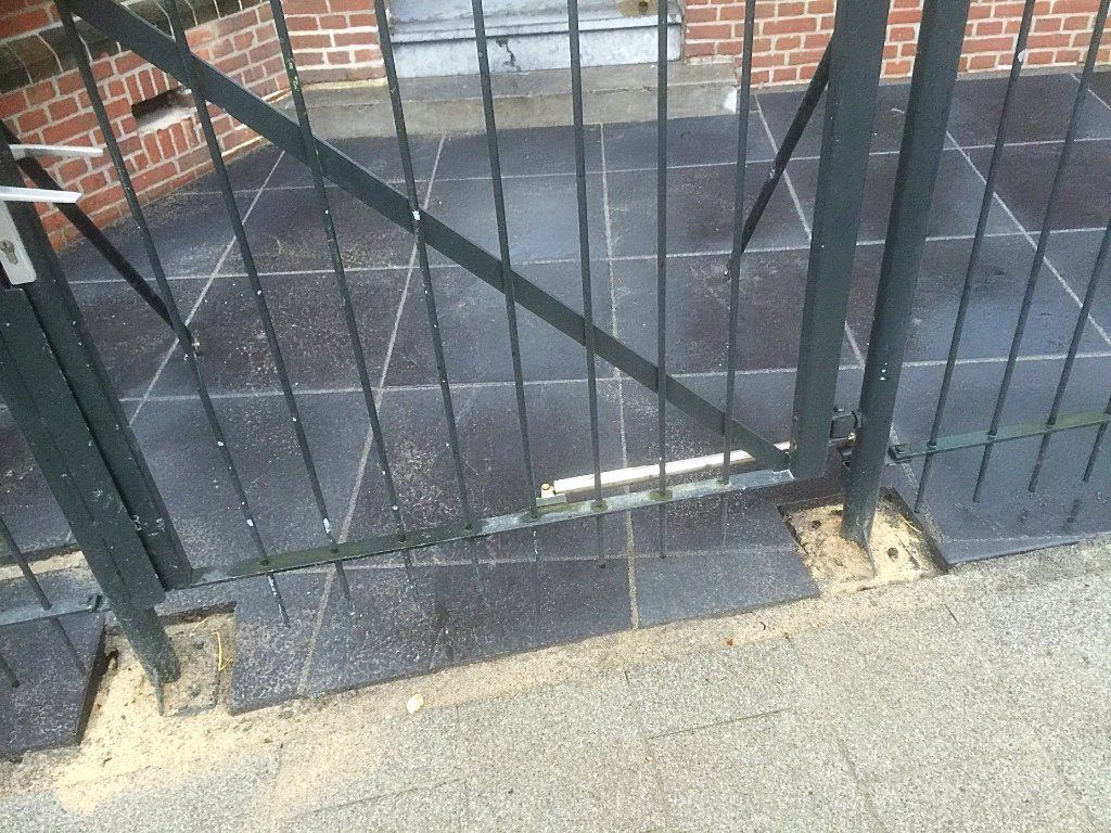 Een zwart metalen hek, bevestigd aan een gebouw, staat een klein beetje open en rust op een betonnen ondergrond.